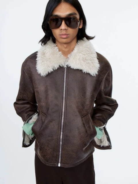 Adult male in a brown aviator jacket with a white fur collar and sunglasses, posing in front of a white backdrop in a studio setting.