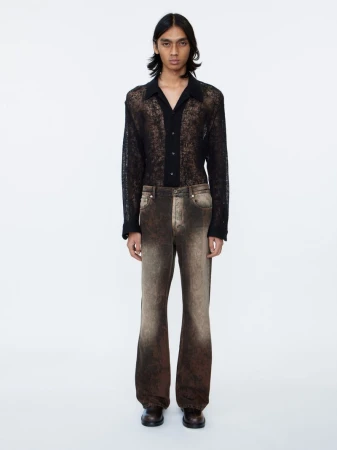 Male model wearing a black sheer button-down shirt and patterned brown trousers against a white seamless background