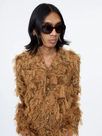 Model wearing dark oval sunglasses and a textured brown fringed shirt against a white backdrop.