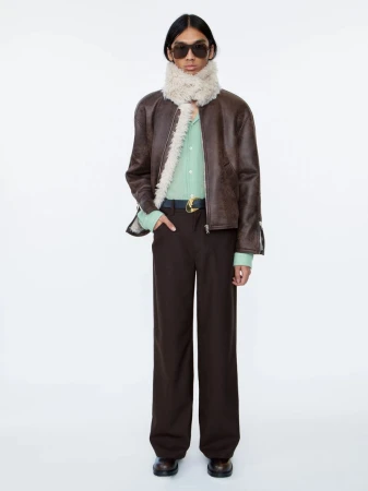 Model wearing a brown leather jacket with shearling trim, dark sunglasses, and dark trousers against a white background