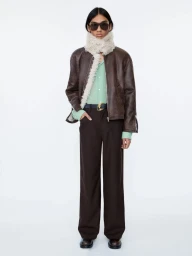 Model wearing a brown leather jacket with shearling trim, dark sunglasses, and dark trousers against a white background