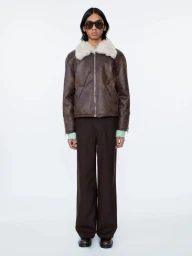Model wearing a brown shearling jacket and dark trousers standing against a white studio seamless background.