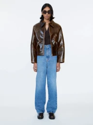 Model wearing a glossy brown leather jacket and blue jeans against a plain light background
