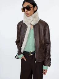 Model wearing dark sunglasses, brown shearling-lined jacket, and green shirt against a white seamless background