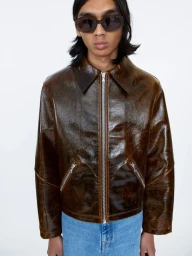 Model wearing dark oval sunglasses, a glossy brown leather jacket, and blue jeans against a plain white background.