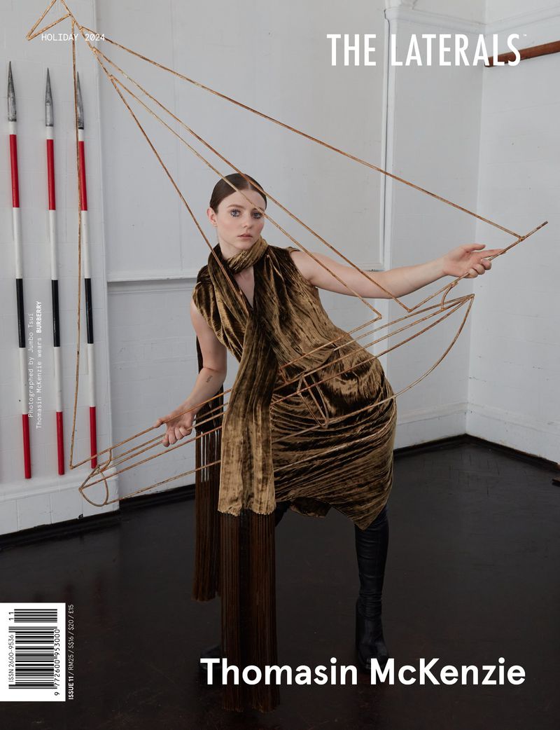 Fashion shot featuring Thomasin McKenzie in a brown dress with geometric wire accents, set against a neutral wall backdrop with spears.