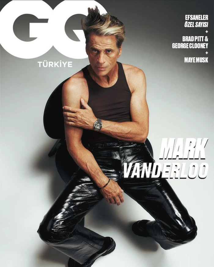 Model Mark Vanderloo wearing a dark tank top and glossy black leather pants seated on a chair against a grey studio backdrop.