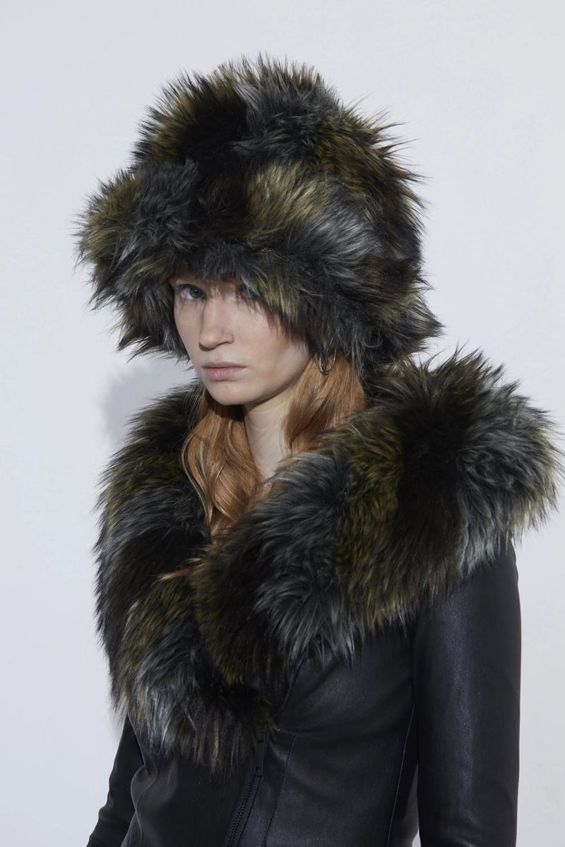 A woman with fair skin and red hair poses in a black leather jacket with fur trim and a matching furry hat against a white backdrop.
