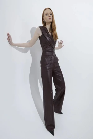 Fashion shot of a red-haired woman in a brown, sleeveless leather jumpsuit. The model poses in front of a white backdrop.