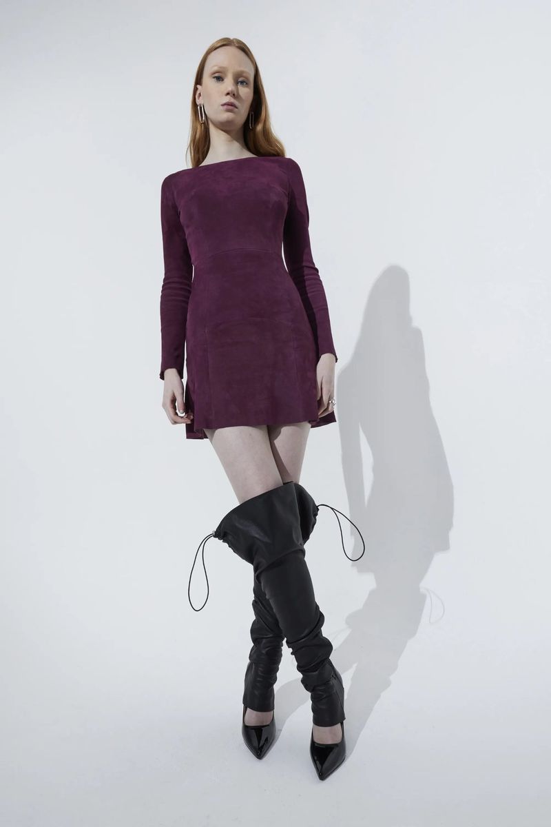 Full-length studio shot of a woman with long red hair wearing a long-sleeve purple mini dress and black over-the-knee heeled boots.