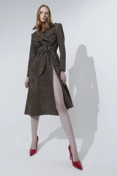 Model with long red hair wearing a dark brown belted coat and red high heels standing against a white backdrop.