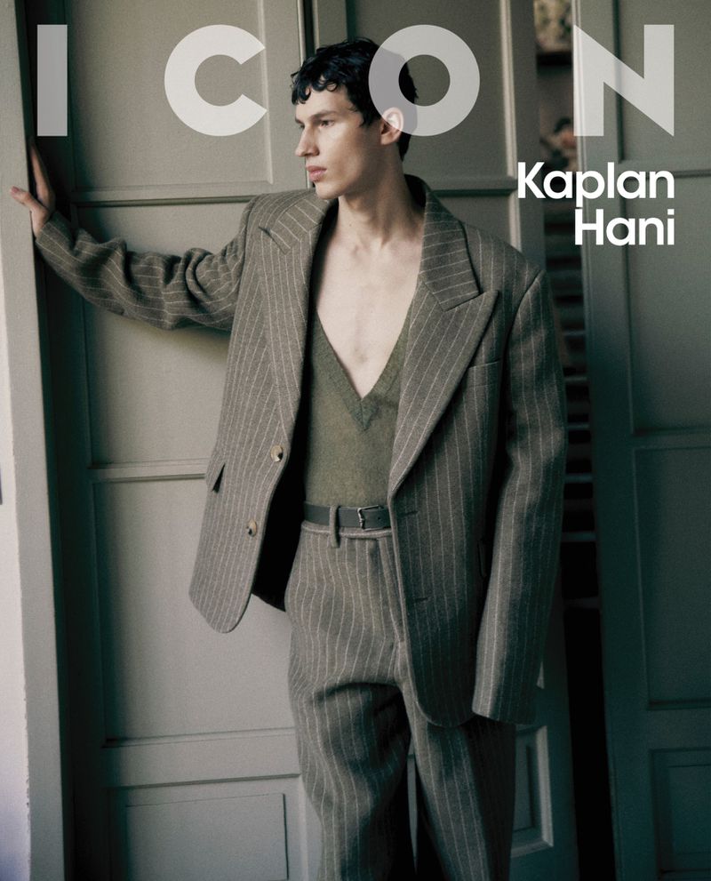 Male fashion model posing in a pinstripe suit jacket and pants near a white door, inside. Editorial-style studio portrait for ICON magazine.