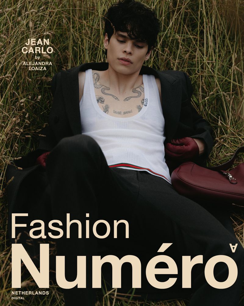 Portrait of a young adult male model wearing a white tank top and dark suit, reclining outdoors among tall grasses for a fashion editorial.