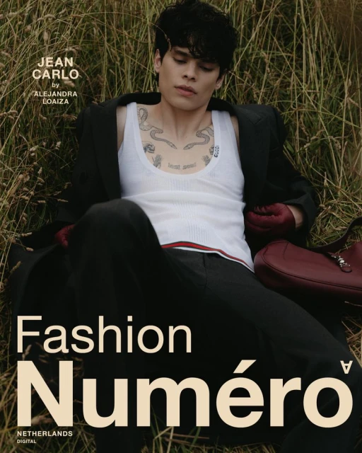 Portrait of a young adult male model wearing a white tank top and dark suit, reclining outdoors among tall grasses for a fashion editorial.