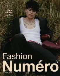 Jean Carlo reclining in tall dry grass wearing a white ribbed tank top, black coat, and red gloves for Numéro Netherlands.