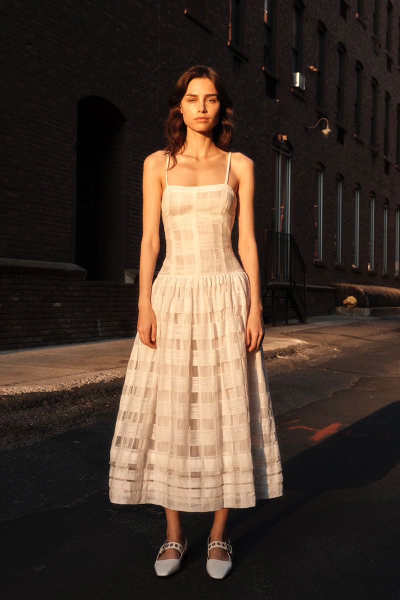 A full-length portrait of a model in a white sundress and white shoes on a city street, posed against a brick building in sunlight.