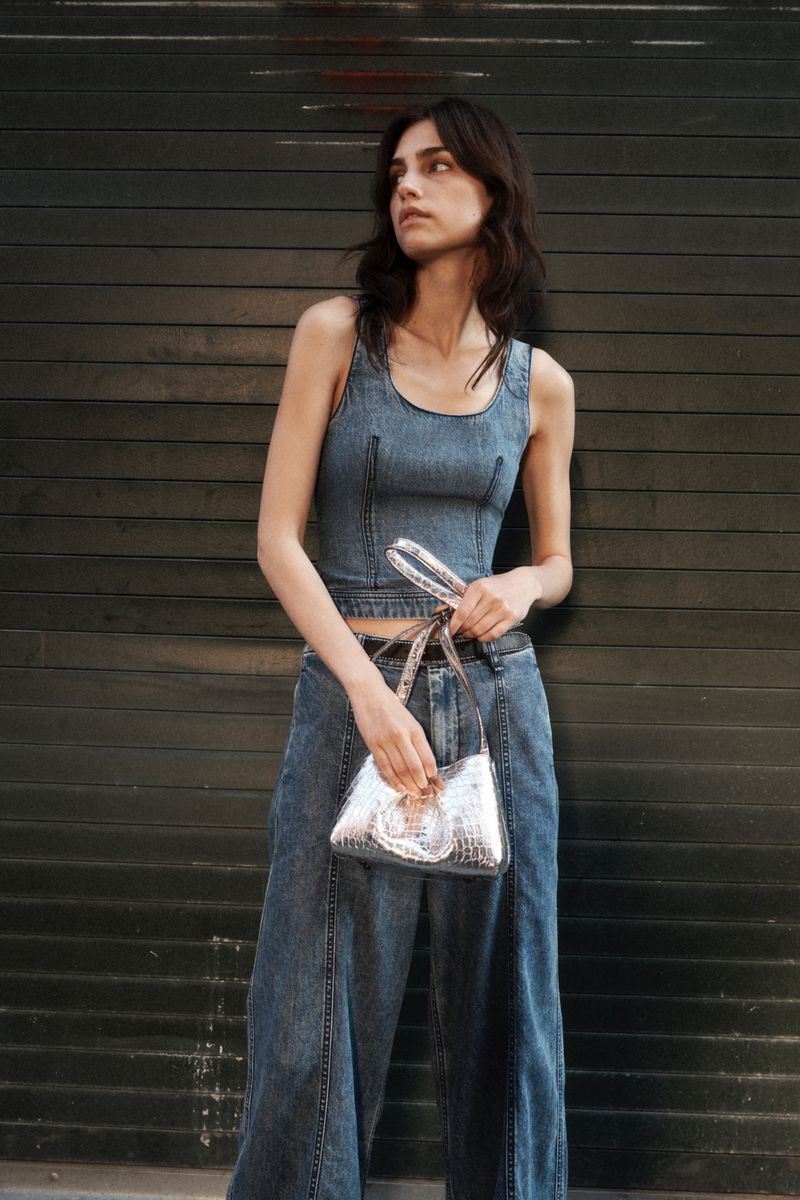 Brunette woman poses in denim crop top and wide-leg jeans with silver purse against a dark background for a fashion shot.