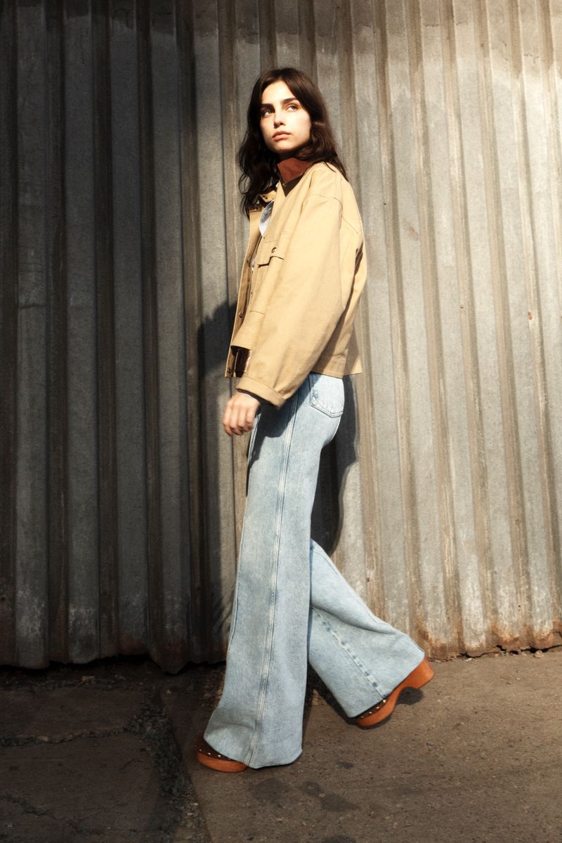 Young woman in a beige jacket and wide-leg jeans poses against a metal wall in an outdoor urban setting with late afternoon light.