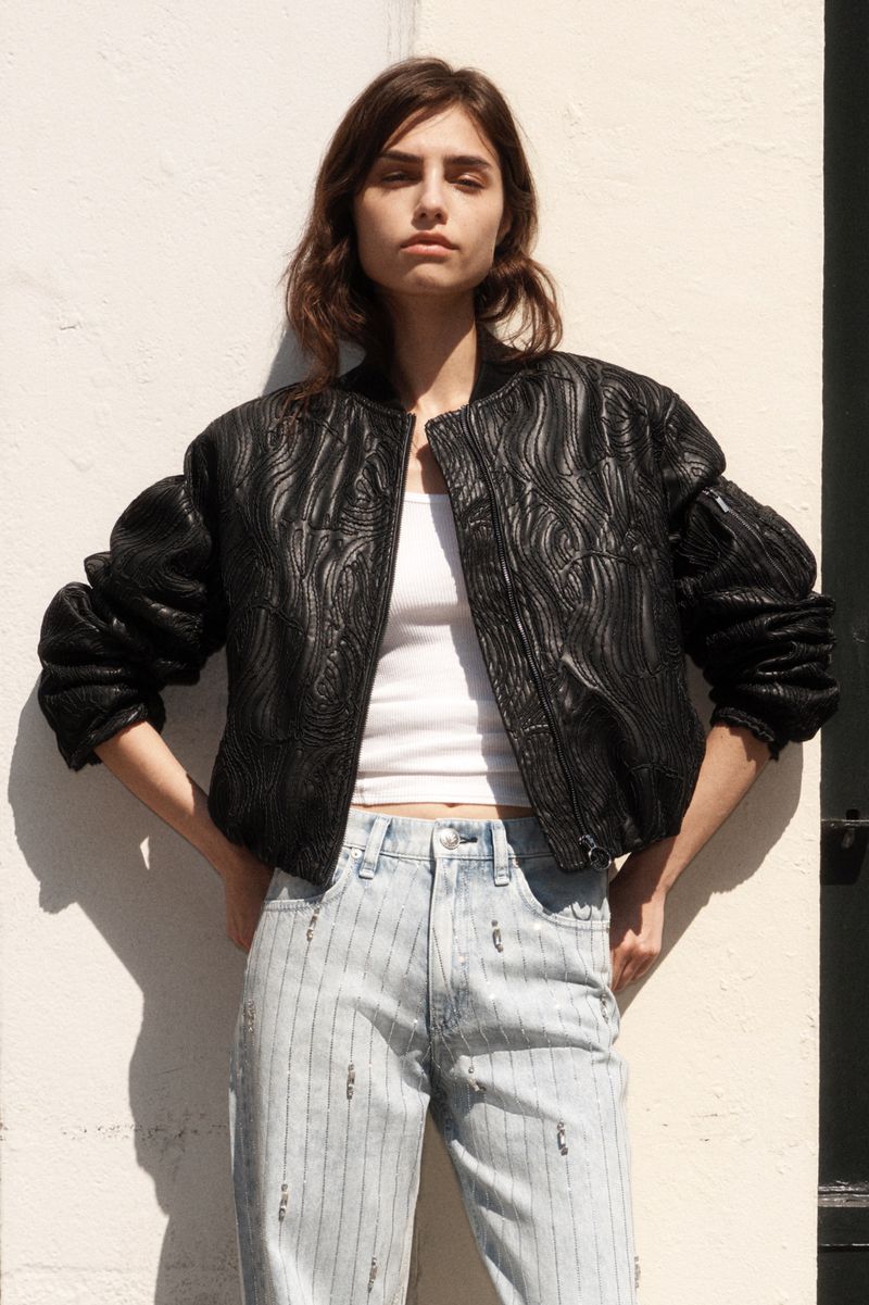 A female fashion model poses outdoors wearing a black patterned bomber jacket, white tank top, and blue pinstripe jeans against a light wall.