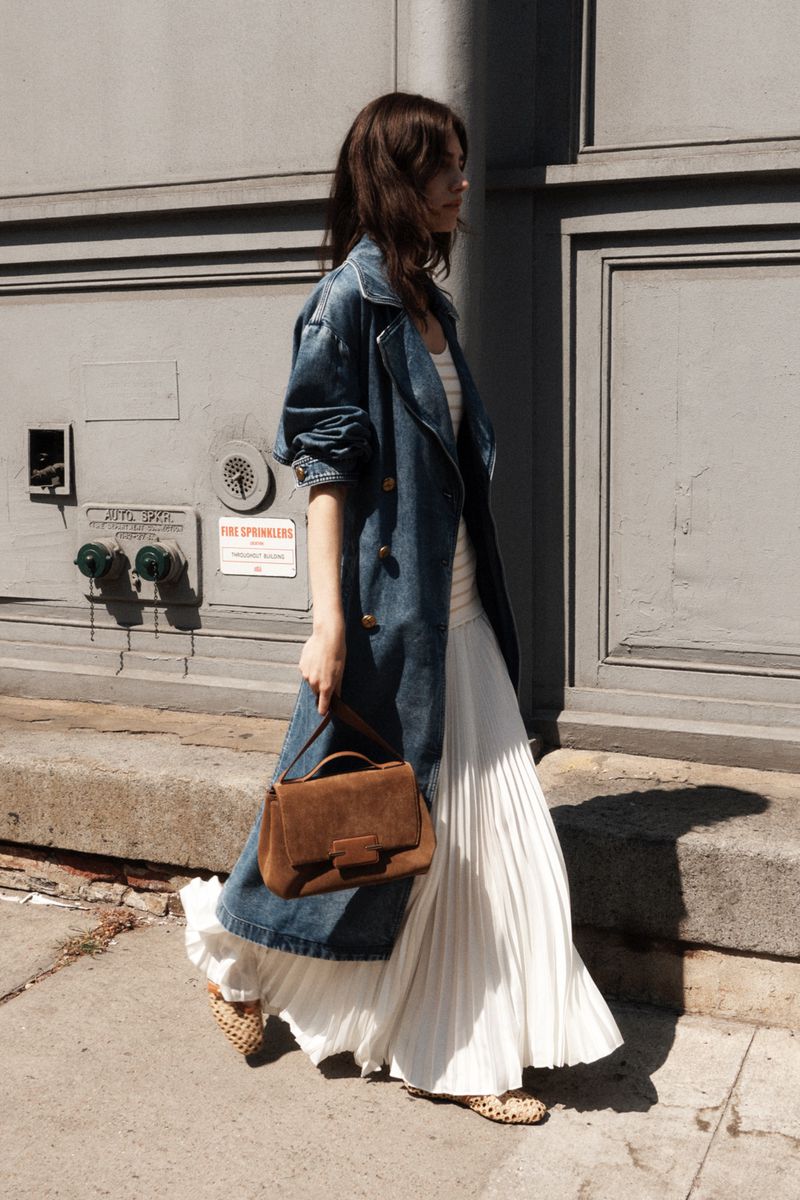A woman walks on a city street wearing a denim trench coat, a white maxi dress, and carrying a brown leather handbag.