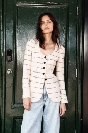 A young woman poses in a cream-colored blazer and light blue jeans against a dark green door, looking confidently at the camera.