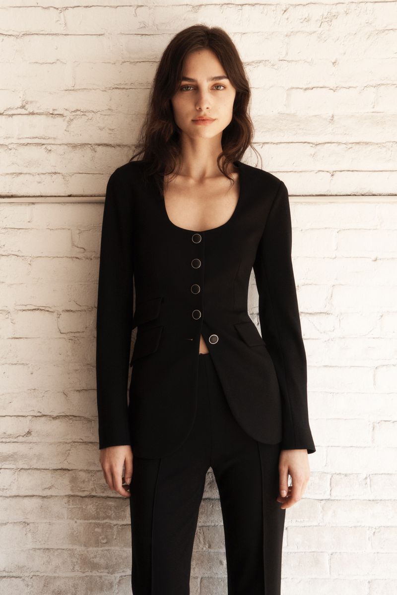 Indoor shot of a woman in a black tailored suit posing against a white brick wall, emphasizing fashion and portrait photography.