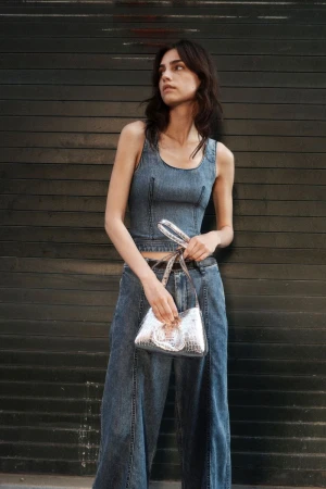 Model in denim top and wide-leg jeans holding a metallic silver handbag against a dark shutter door