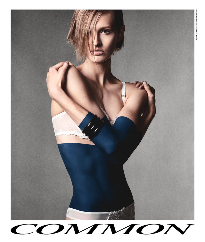 Fashion editorial shot of a woman with short blonde hair wearing a white bra and blue arm sleeves against a gray backdrop.