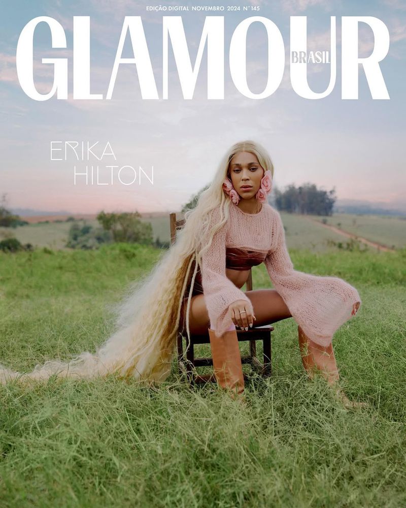 Glamour Brasil magazine cover featuring Erika Hilton with long blonde hair, wearing a pink knit top, sitting on a chair in a field.