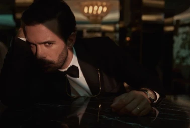 Man with dark hair and a beard wearing a black tuxedo and bow tie, leaning his head on his hand against a dark marble table.