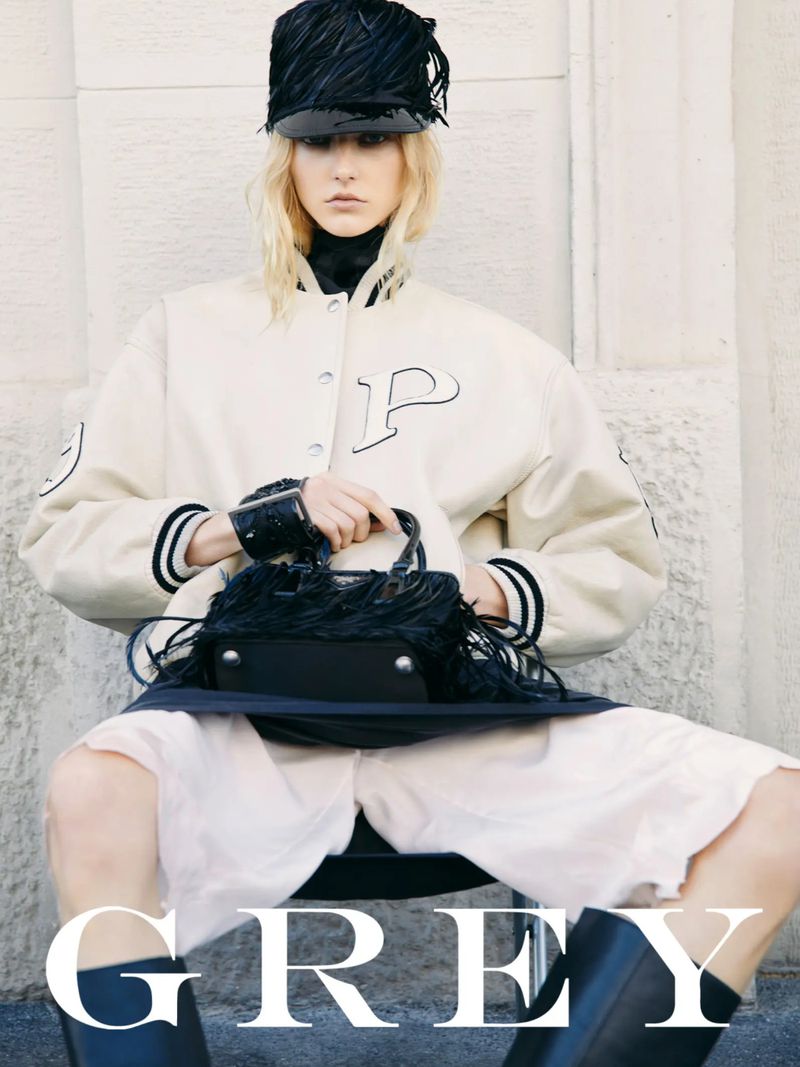 A fashionable fair-skinned woman in a letterman jacket, hat, and boots, seated and holding a fringed handbag against a neutral backdrop.