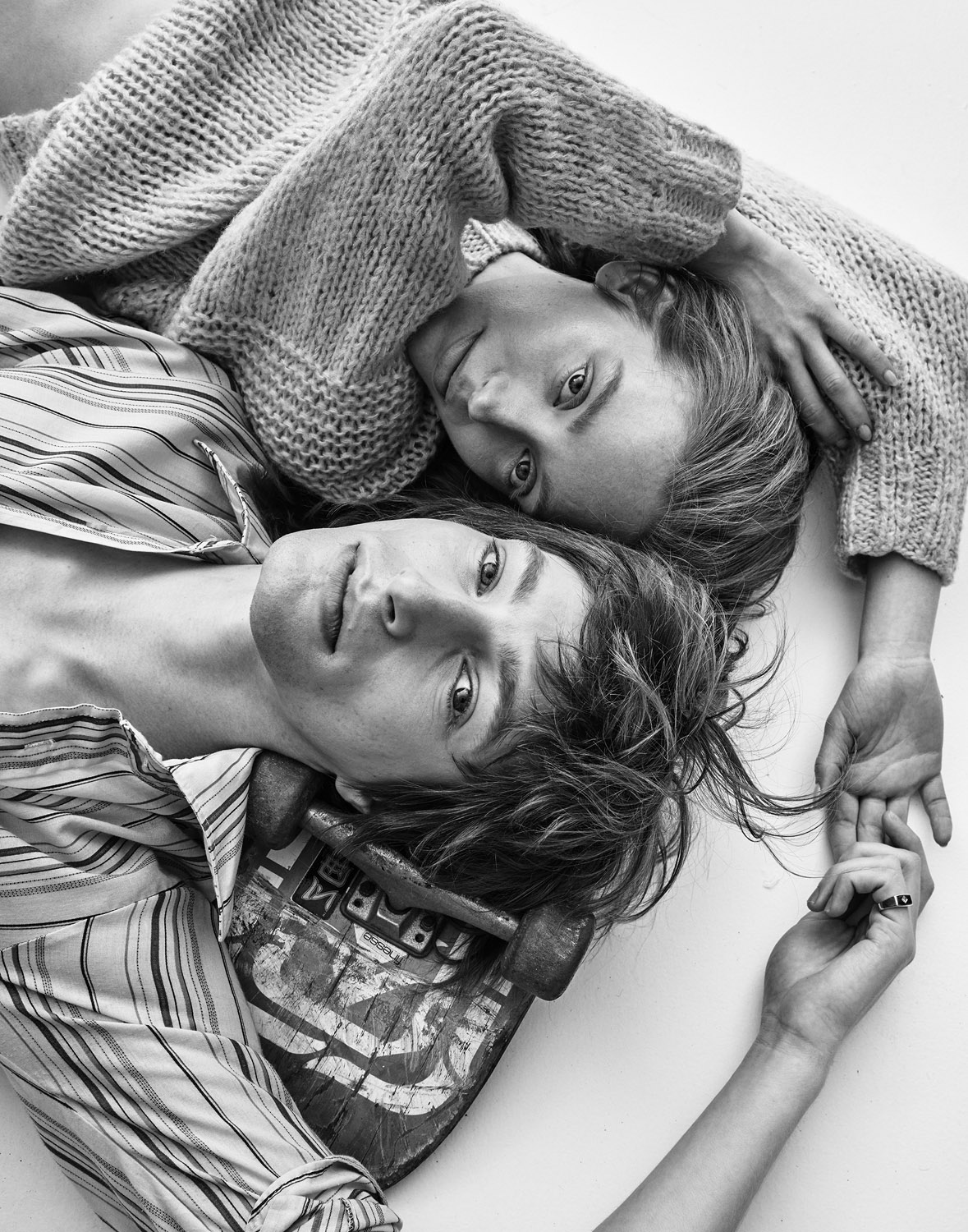 Black-and-white studio shot of a couple lying close together, looking directly at the camera, the man laying on a skateboard.