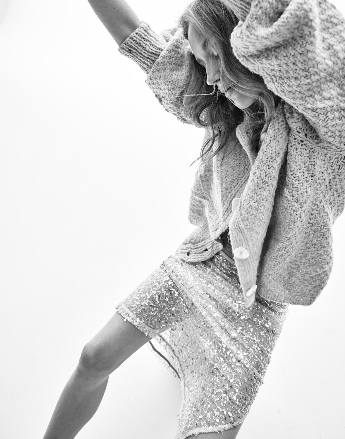 Black and white studio shot of a blond female model in a knit cardigan and sparkling mini skirt, posing with her hands on her head.