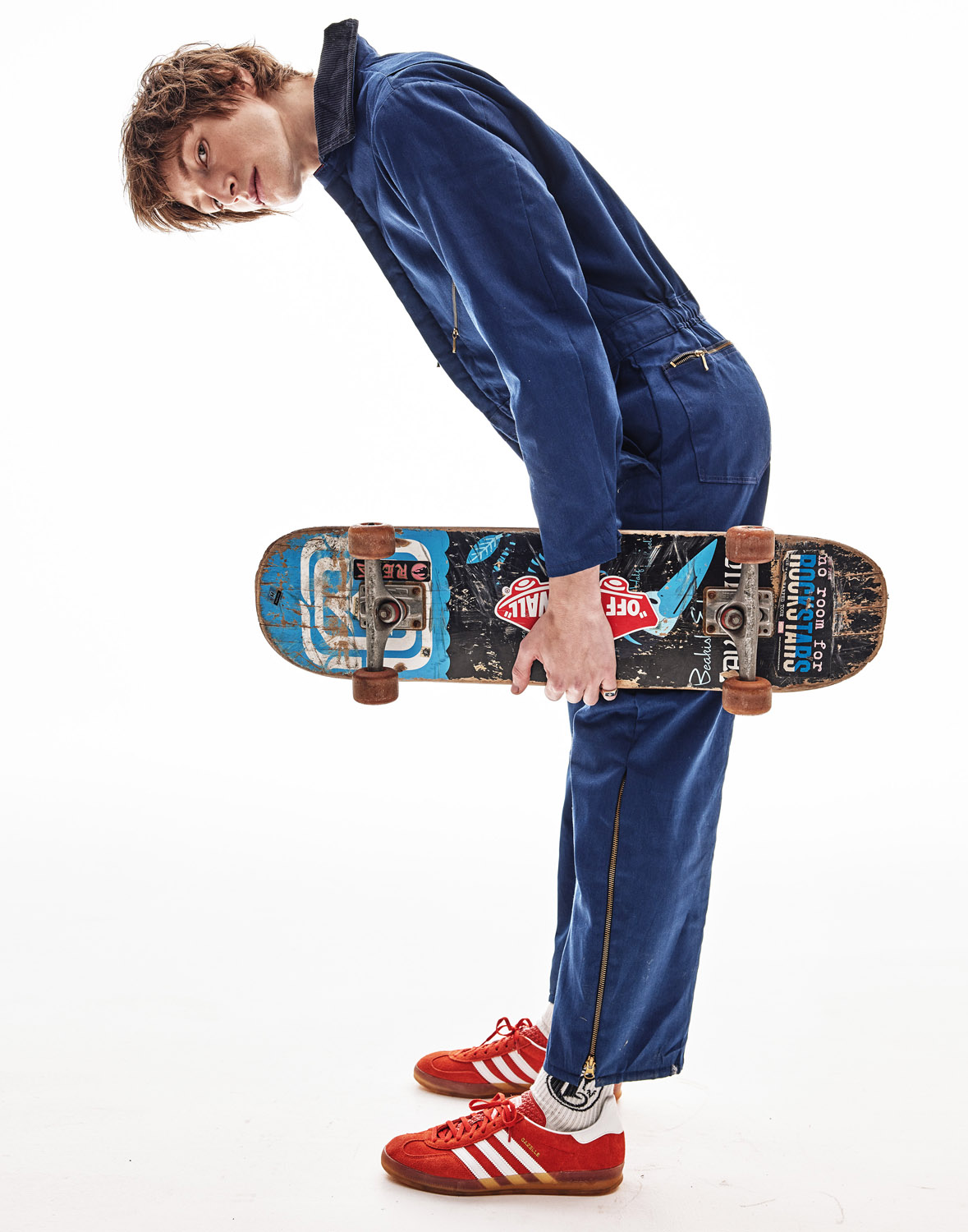 Young adult man in a blue jumpsuit poses with a skateboard in a studio setting. Red Adidas sneakers add a pop of color.