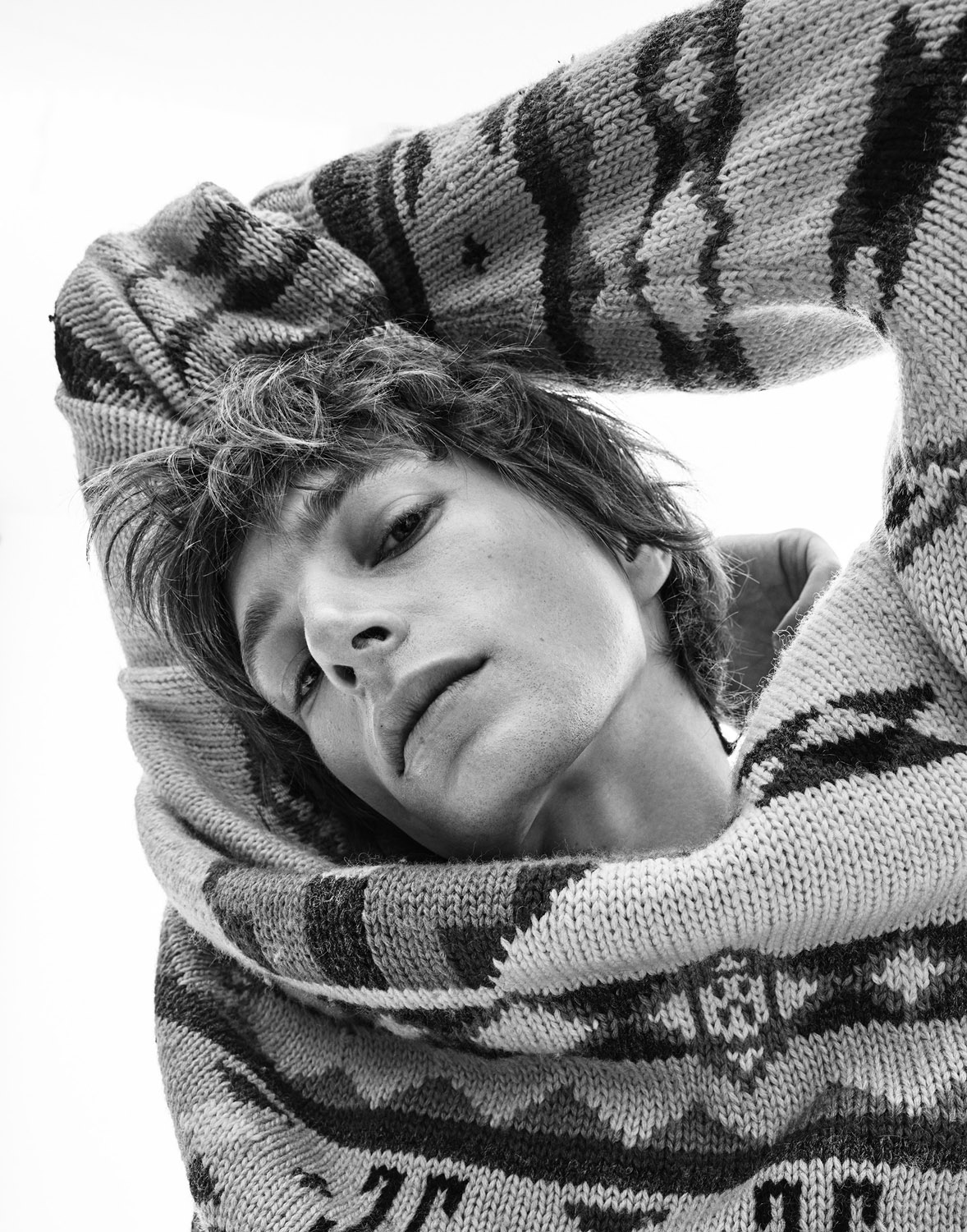 Monochrome portrait of an adult man with medium-length hair in patterned sweater, posing against a plain white backdrop in studio setting.
