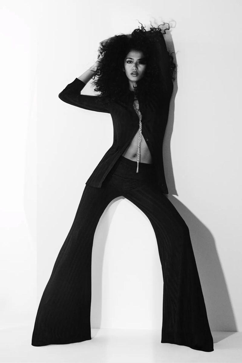 Black and white portrait of a woman in a suit posing against a stark white background with dramatic lighting and large curly hair.