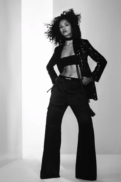 A woman poses in black crop top, blazer, and wide-leg pants in a black and white studio fashion shot with a light background.