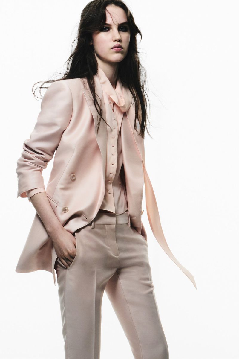 Woman in a light pink suit poses in a studio. The blazer is double-breasted with a matching blouse, and trousers, against a white backdrop.