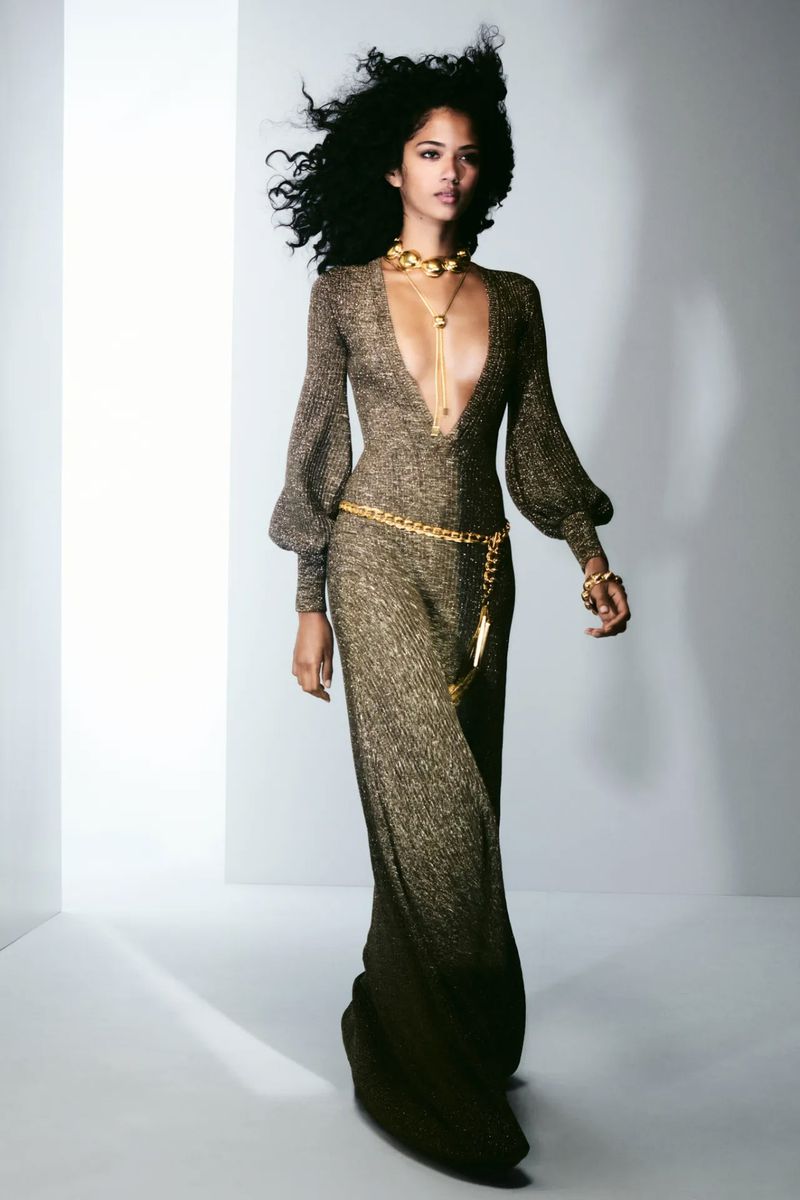 Elegant adult female with dark curly hair in a shimmering long gown with gold jewelry in a bright, neutral studio setting.
