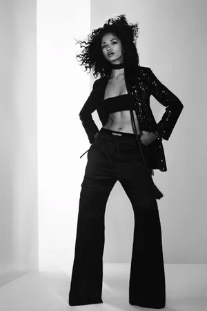 Model with curly hair wearing a black sequined jacket, bandeau top, and trousers against a light studio backdrop