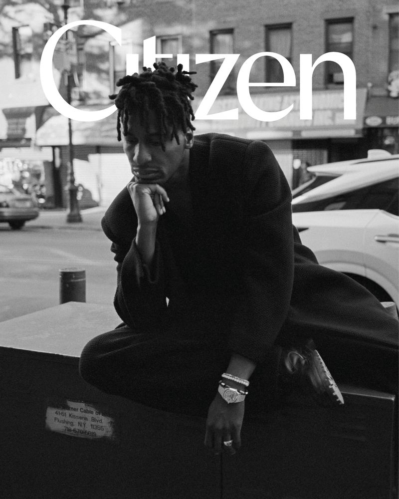 Black and white portrait of an adult man with dreadlocks in an urban setting, sitting with his hand to his face, wearing a dark sweater.