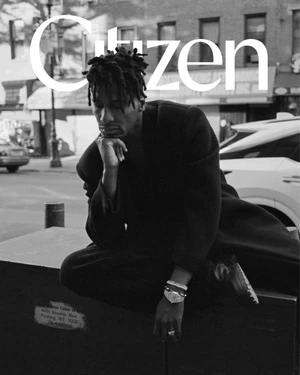 Black and white portrait of Jon Batiste seated on a street structure wearing a dark coat and bracelets, with city street background.