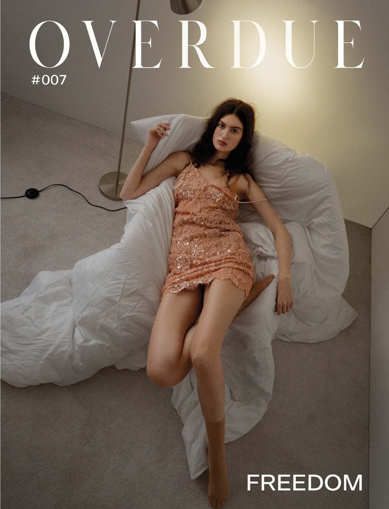 Brunette woman in a sequined dress reclines on a white bed in a studio setting with a floor lamp for a fashion magazine cover.