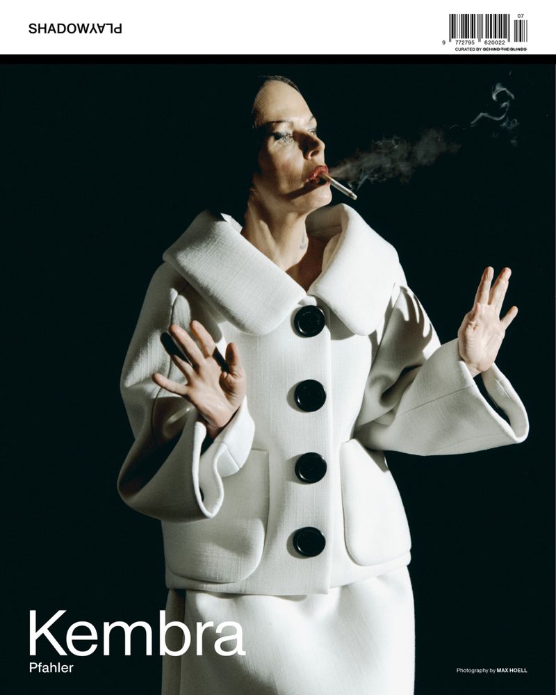 Woman in a white suit with black buttons smoking a cigarette against a dark background for a fashion magazine cover.