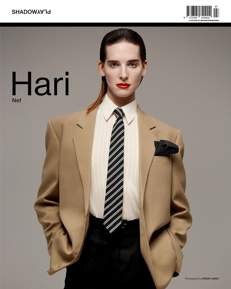 Portrait of Hari Nef wearing a beige blazer, striped shirt, and black tie, posing in front of a neutral gray background for a fashion editorial.