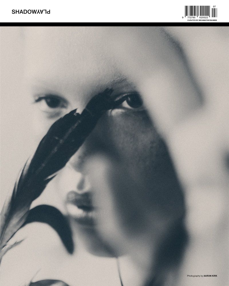 Monochromatic close-up studio portrait of a fair-skinned woman with a feather covering one eye and blurred hand in front of face.