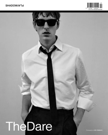 Black-and-white fashion shot of a young adult man in a white shirt, black tie, and sunglasses, standing with his hands in his pockets.