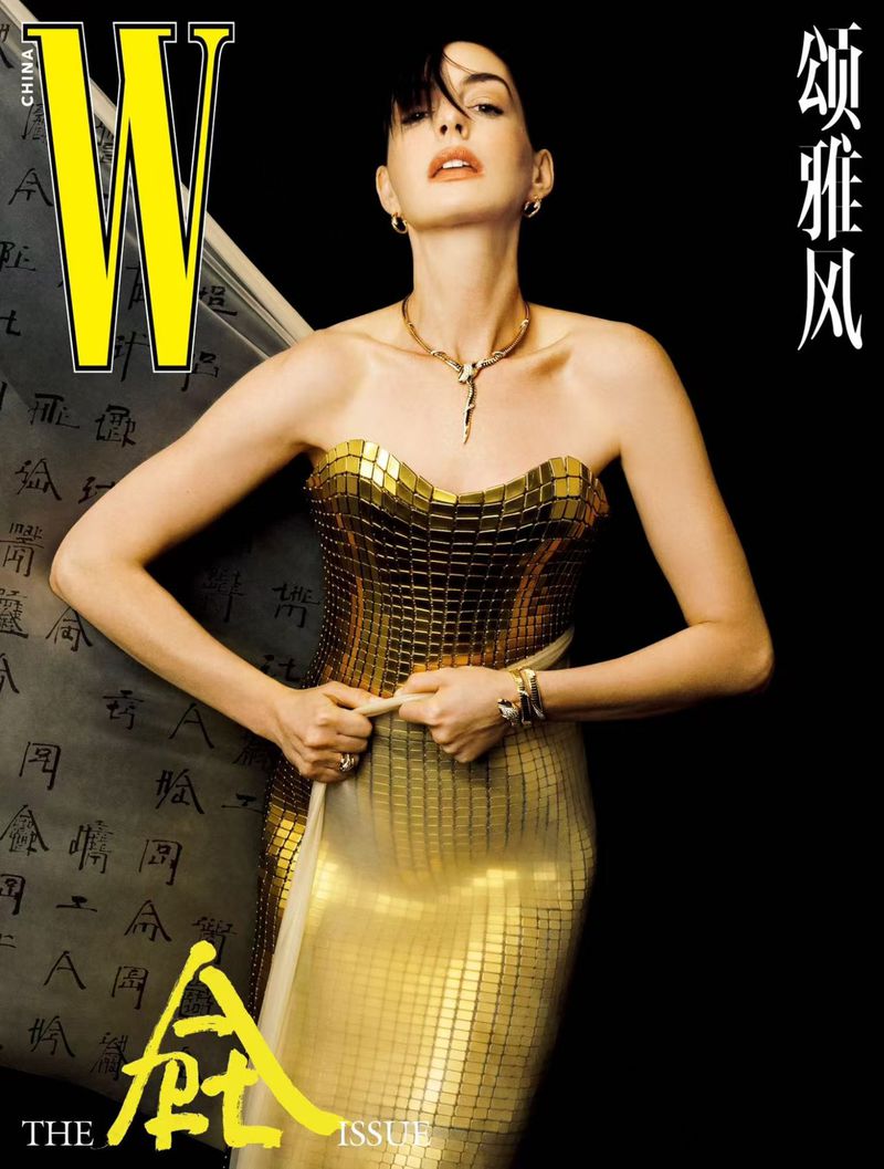 Anne Hathaway in a gold strapless dress for W Magazine China, posing against a black background with a scroll with East Asian calligraphy.