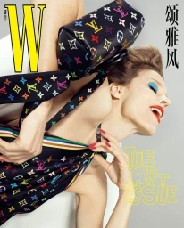 Model with blue eyeshadow and red lipstick holding a colorful monogram handbag against a neutral grey studio backdrop.