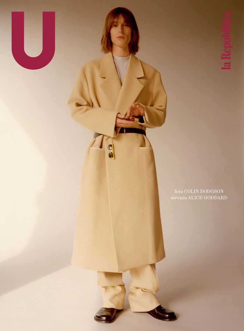 Fashion editorial shot of a slender male model wearing a beige overcoat and trousers, posing indoors against a neutral-toned backdrop.